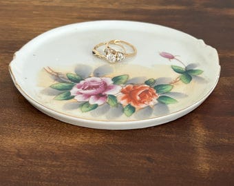 May include: A white ceramic trinket dish with a floral design featuring pink, orange, and green roses. The dish has a gold rim and is perfect for holding jewelry or other small items.