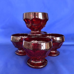 May include: Four ruby red, vintage-style dessert glasses with a gold rim. The glasses have a faceted design and are arranged against a blue background. The glasses are likely made of glass.