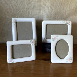 May include: A set of four white ceramic picture frames with rounded corners and a delicate floral pattern featuring small pink flowers and green leaves. The collection includes two rectangular frames in portrait and landscape, one horizontal frame, and one oval frame.
