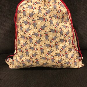 May include: A beige drawstring bag with a red drawstring. The bag is covered in a pattern of gray sock monkeys with red hats.