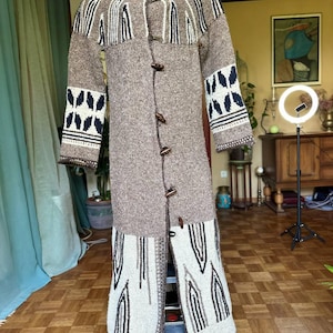 Pode incluir: Um casaco comprido de malha castanha com gola alta e fechos de alternância de madeira. O casaco apresenta padrões geométricos em branco, azul marinho e castanho nos ombros, mangas e bainha inferior. O casaco é exibido num cabide de metal.