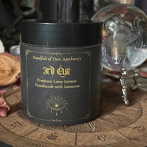 Puede incluir: Un tarro negro de incienso suelto etiquetado como "Handfuls of Dust Apothecary 3rd Eye Premium Loose Incense Handmade with Intention". El tarro está sobre una superficie de madera con un diseño circular y un orbe de vidrio sobre un soporte.