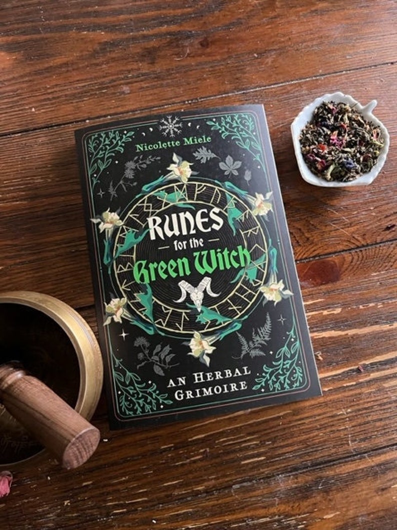 May include: A black book with a green and white illustration of runes and flowers. The title is "Runes for the Green Witch: An Herbal Grimoire" by Nicolette Miele.