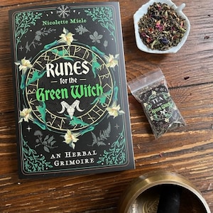 May include: A black book with a green and gold design featuring the title "Runes for the Green Witch: An Herbal Grimoire" by Nicolette Miele. The book is on a wooden table with a small white bowl of tea, a small bag of tea, and a brass singing bowl.