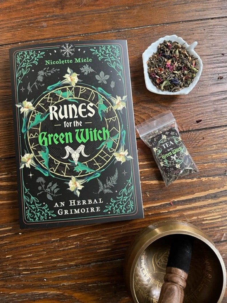 May include: A black book with a green and gold design featuring the title "Runes for the Green Witch: An Herbal Grimoire" by Nicolette Miele. The book is on a wooden table with a small white bowl of tea, a small bag of tea, and a brass singing bowl.