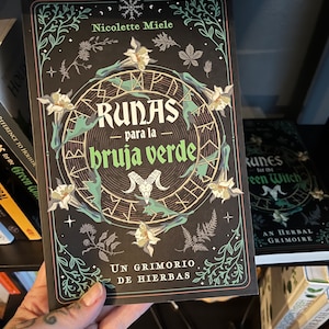 May include: A book titled "Runas para la bruja verde: Un grimorio de hierbas" by Nicolette Miele. The cover features a black background with a circular design of runes, a skull, and floral illustrations in shades of green, white, and gold.