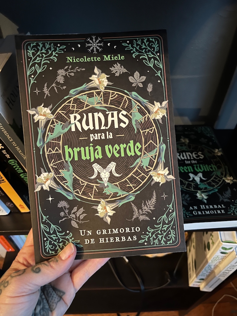 May include: A book titled "Runas para la bruja verde: Un grimorio de hierbas" by Nicolette Miele. The cover features a black background with a circular design of runes, a skull, and floral illustrations in shades of green, white, and gold.