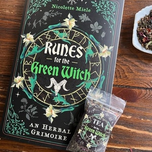 May include: A black book with a green and white floral design and the title "Runes for the Green Witch: An Herbal Grimoire" by Nicolette Miele. A small bag of tea with the text "Tea for the Green Witch" is in the foreground.