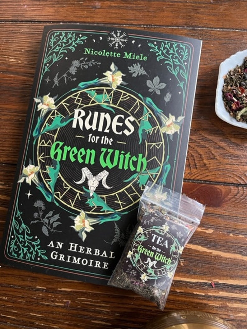 May include: A black book with a green and white floral design and the title "Runes for the Green Witch: An Herbal Grimoire" by Nicolette Miele. A small bag of tea with the text "Tea for the Green Witch" is in the foreground.