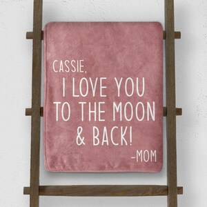 May include: A dusty rose blanket with the text "Cassie, I love you to the moon & back! -Mom" in white lettering. The blanket is draped over a wooden ladder.