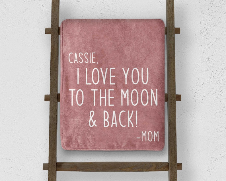 May include: A dusty rose blanket with the text "Cassie, I love you to the moon & back! -Mom" in white lettering. The blanket is draped over a wooden ladder.