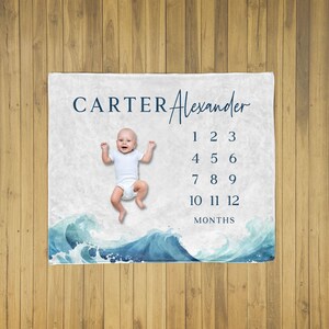 May include: A personalized baby milestone blanket with the name "Carter Alexander" in navy blue script. The blanket features a watercolor wave design at the bottom and a space to track the baby's age in months, from 1 to 12. A baby is lying on the blanket.