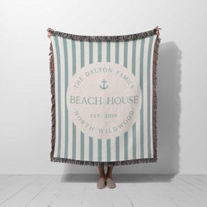 May include: A woven throw blanket with a white and teal striped pattern. The blanket features a circular logo with the text "The Dalton Family Beach House Est. 2019 North Wildwood".