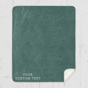 May include: A teal-colored blanket with a textured surface and a white sherpa fleece edge. The blanket has the text "YOUR CUSTOM TEXT" printed in white at the bottom. The blanket is displayed on a white wooden surface.