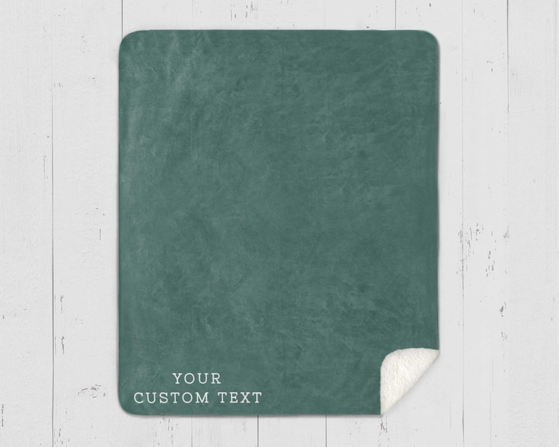 May include: A teal-colored blanket with a textured surface and a white sherpa fleece edge. The blanket has the text "YOUR CUSTOM TEXT" printed in white at the bottom. The blanket is displayed on a white wooden surface.