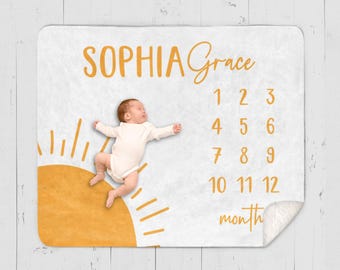 May include: A white baby blanket with a yellow sun design and numbers 1-12 for tracking a baby's monthly growth. The text "Sophia Grace" and "month" are printed on the blanket. A baby is lying on the blanket.