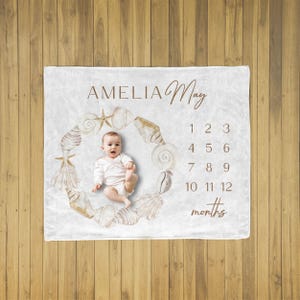 May include: A white baby milestone blanket with the name "Amelia May" in brown script. A wreath of seashells and starfish surrounds a baby. Numbers 1-12 and the word "months" are also printed on the blanket.