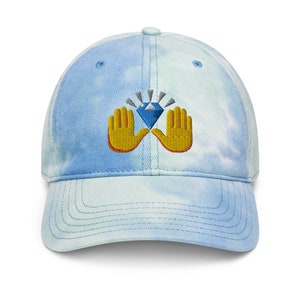 May include: A baseball cap with a tie-dye pattern in shades of blue and white. The cap features an embroidered design of a blue diamond held by two yellow hands.