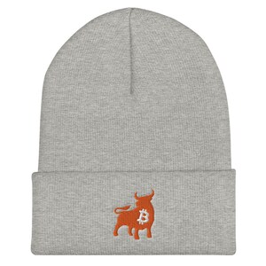 May include: A gray beanie featuring an embroidered orange bull with a Bitcoin symbol. The beanie is made of a soft, knitted material and has a folded cuff. The bull graphic is located on the lower front of the hat.
