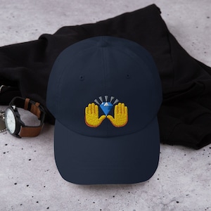 May include: Navy blue baseball cap with an embroidered design of a blue diamond held by two yellow hands. The cap is made of a soft material and has a curved brim. A stylish accessory for everyday wear.