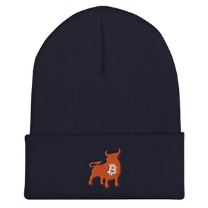 May include: A navy blue beanie featuring an embroidered orange bull with a Bitcoin symbol. The beanie is made of a soft, ribbed knit material. The bull graphic is located on the front of the folded brim.