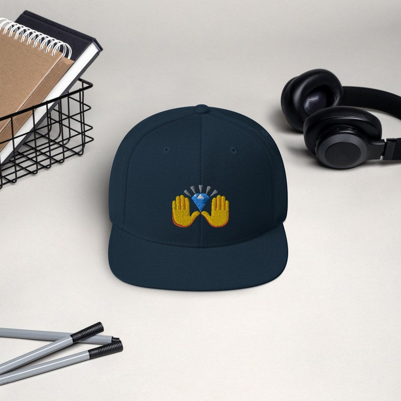 Pu&ograve; includere: Cappellino da baseball blu navy con visiera piatta. Il cappellino presenta un design ricamato di due mani gialle che tengono un diamante blu con luce radiante. Un accessorio alla moda.