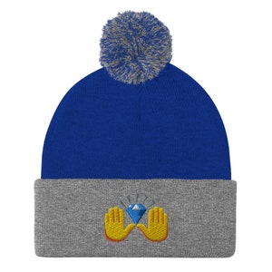 May include: A blue beanie with a gray brim and a gray pom-pom. The front of the beanie features an embroidered design of a blue diamond held by two yellow hands.