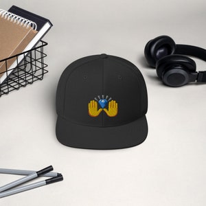 Pu&ograve; includere: Cappellino da baseball nero a tesa piatta con un design ricamato. Il design presenta un diamante blu tenuto da due mani gialle con contorni arancioni. Uno stile classico, adatto all'uso quotidiano.