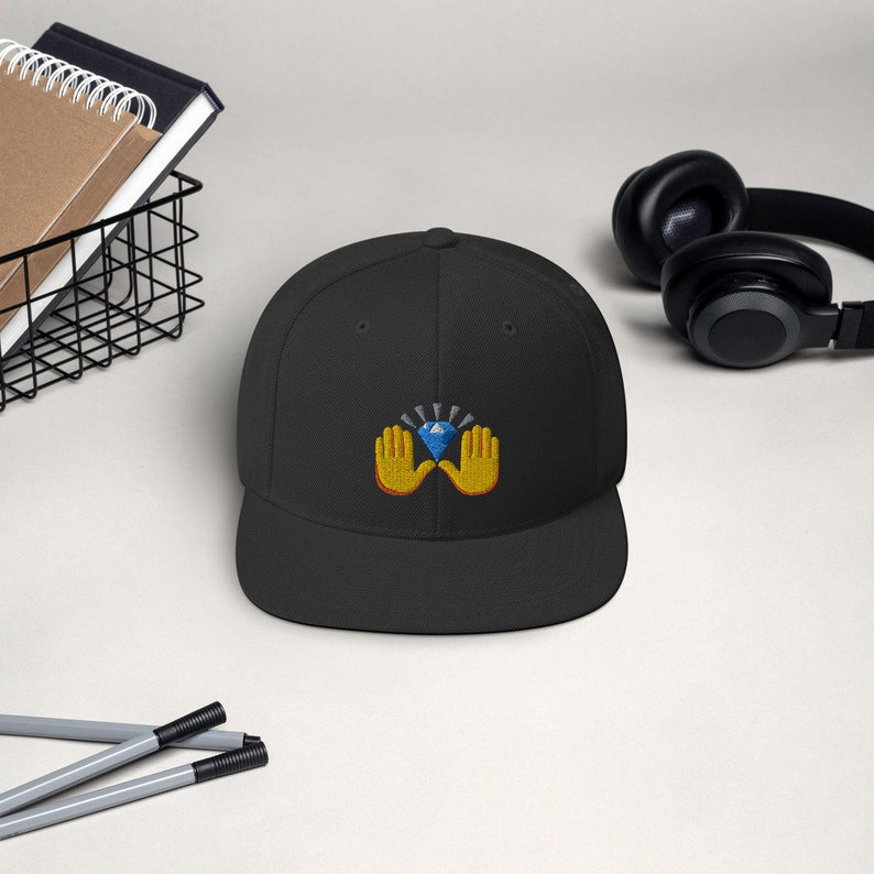 Pu&ograve; includere: Cappellino da baseball nero a tesa piatta con un design ricamato. Il design presenta un diamante blu tenuto da due mani gialle con contorni arancioni. Uno stile classico, adatto all'uso quotidiano.