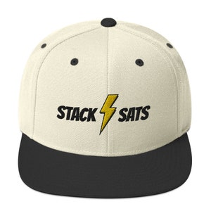 Puede incluir: Gorra de béisbol de ala plana color crema con visera negra. La gorra presenta el texto bordado "STACK SATS" en negro con un rayo amarillo.
