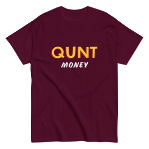 May include: A maroon t-shirt with the word "QUNT" in yellow with a red shadow, and the word "MONEY" in white. The shirt is made of a soft material and has a crew neck.