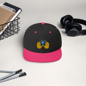 Pu&ograve; includere: Un cappellino da baseball nero con visiera e bottone superiore rosa acceso. Il cappellino presenta un design ricamato di mani gialle che tengono un diamante blu. Un accessorio elegante per l'uso quotidiano.