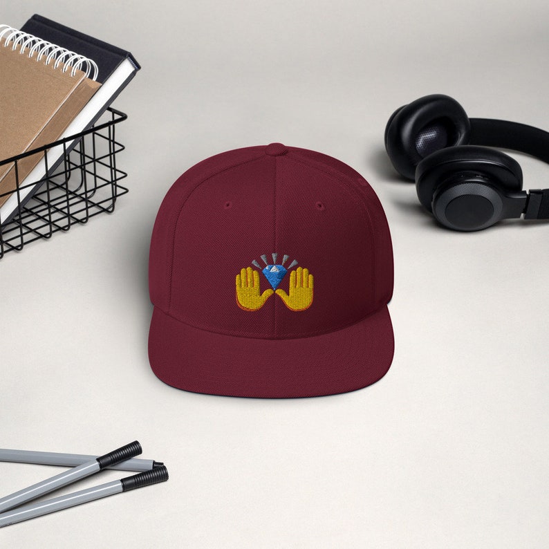 Pu&ograve; includere: Cappellino da baseball bordeaux con visiera piatta. Il cappellino presenta un design ricamato di due mani gialle che sorreggono un diamante blu con raggi di luce radianti. Il cappellino &egrave; in stile snapback.