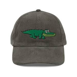 Könnte beinhalten: Graue Cord-Baseballkappe mit einem gestickten grünen Alligatorendesign. Der Alligator hat einen grünen Körper, einen weißen Bauch und einen grün-weißen Mund. Die Kappe hat einen gebogenen Schirm und einen verstellbaren Riemen.