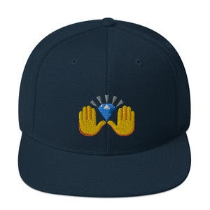 Pu&ograve; includere: Un cappellino da baseball blu navy con visiera piatta. Il cappellino presenta un disegno ricamato di un diamante blu tenuto da due mani gialle con contorni arancioni. Il diamante ha raggi bianchi e argentati.