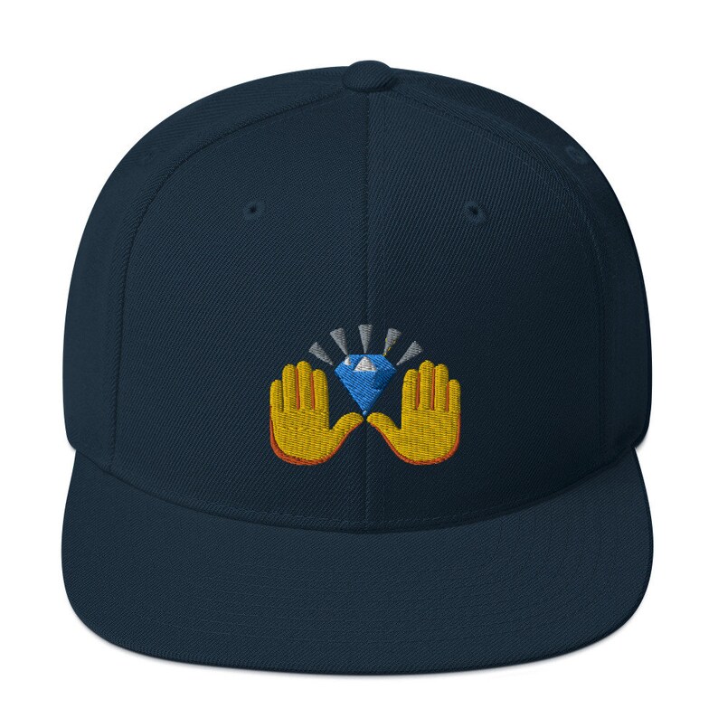 Pu&ograve; includere: Un cappellino da baseball blu navy con visiera piatta. Il cappellino presenta un disegno ricamato di un diamante blu tenuto da due mani gialle con contorni arancioni. Il diamante ha raggi bianchi e argentati.