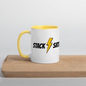 Puede incluir: Taza de cerámica blanca con interior y asa amarillos. La taza presenta el texto "STACK SATS" en negro con un rayo amarillo. La taza está sobre una superficie de madera.
