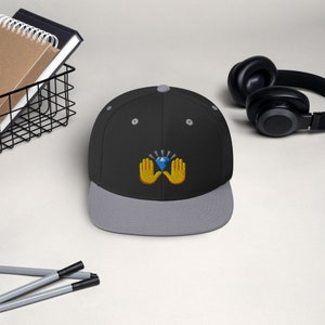 Pu&ograve; includere: Un cappellino da baseball nero e grigio con visiera piatta. Il cappellino presenta un design ricamato di due mani gialle che sorreggono un diamante blu con linee radianti. Il cappellino ha una vestibilit&agrave; strutturata e una chiusura a scatto.