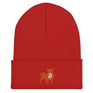 May include: A red beanie with a folded brim. The beanie features an embroidered design of a bull with a Bitcoin symbol in the center. The bull is orange, and the Bitcoin symbol is silver.