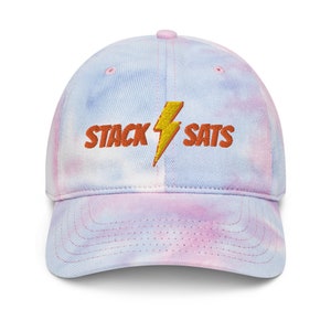 Puede incluir: Gorra de béisbol teñida con tinte anudado en tonos rosa, azul y blanco. La gorra presenta el texto bordado "STACK SATS" con un rayo amarillo.