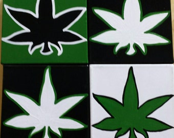 Puede incluir: Cuatro pequeños lienzos cuadrados con un diseño de hoja de marihuana verde, negro y blanco pintado en cada uno. Las hojas están delineadas en negro.