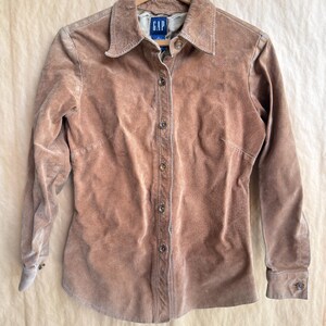 May include: A tan suede button-down shirt with long sleeves, a pointed collar, and a front button placket. The garment features a Gap brand label at the inner neckline and is displayed on a wooden hanger against a neutral backdrop.