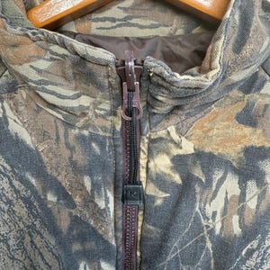 May include: Close-up of a camouflage jacket with a brown zipper. The jacket features a woodland camouflage pattern in shades of brown, green, and tan. The zipper is dark brown, and the collar is visible. The jacket is hanging on a wooden hanger.