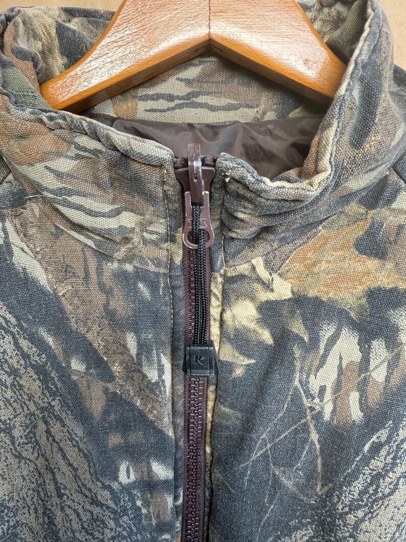 May include: Close-up of a camouflage jacket with a brown zipper. The jacket features a woodland camouflage pattern in shades of brown, green, and tan. The zipper is dark brown, and the collar is visible. The jacket is hanging on a wooden hanger.
