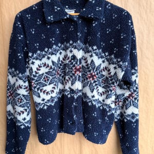 May include: A navy blue fleece jacket with a white and red snowflake pattern. The jacket has a collared neckline and button-up closure. The jacket is hanging on a wooden hanger.