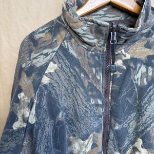 May include: A camouflage zip-up jacket in shades of brown, gray, and green. The jacket features a high collar and a full-length zipper. The fabric has a pattern of leaves and branches.