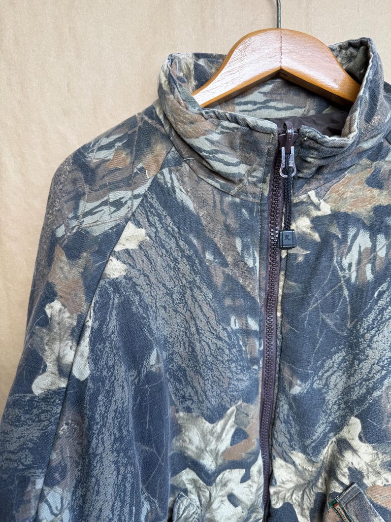 May include: A camouflage zip-up jacket in shades of brown, gray, and green. The jacket features a high collar and a full-length zipper. The fabric has a pattern of leaves and branches.