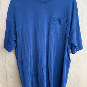 May include: A solid blue t-shirt with a chest pocket. The short-sleeved shirt is made of a soft material and hangs on a wooden hanger. The shirt has a relaxed fit and a crew neck.