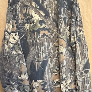 K&ouml;nnte beinhalten: Eine Tarnjacke in Braun-, Gr&uuml;n- und Graut&ouml;nen mit Knopfleiste und Kragen. Das Muster der Jacke ahmt eine Waldumgebung nach, mit detaillierten Darstellungen von Bl&auml;ttern und &Auml;sten.