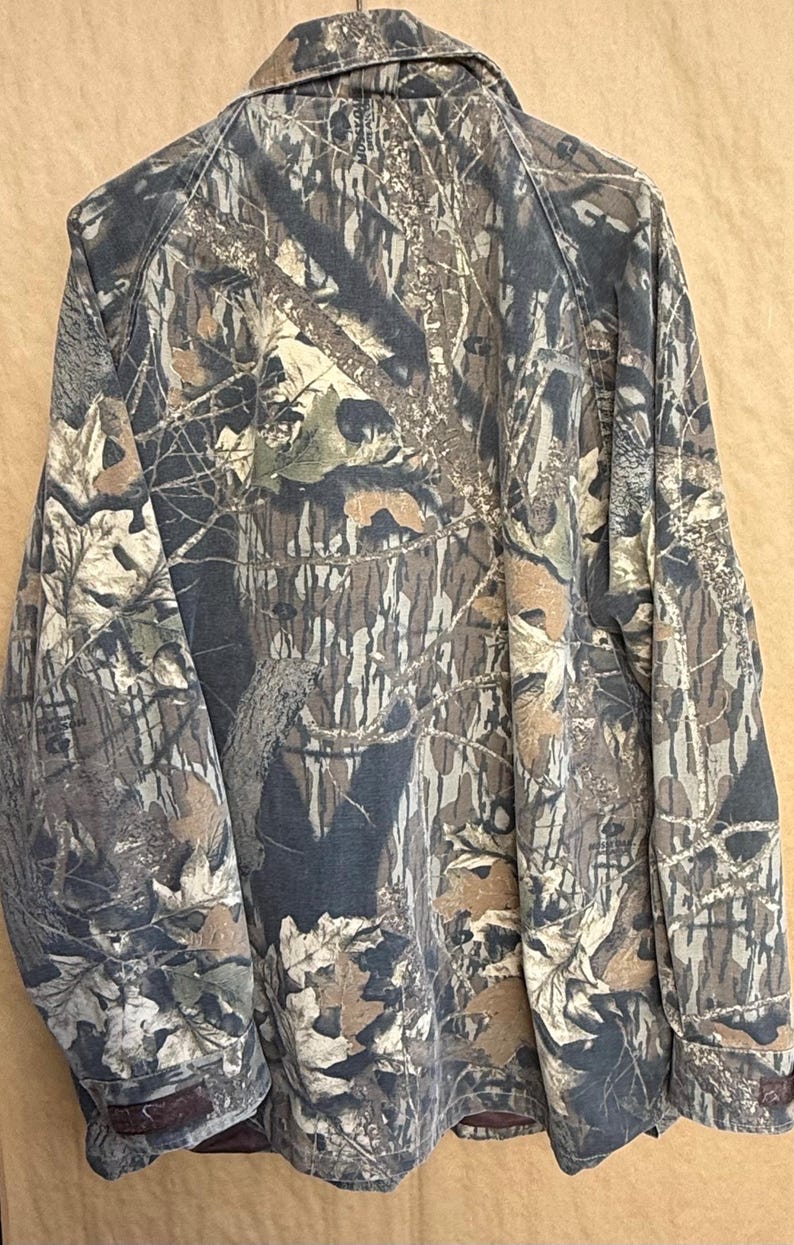 K&ouml;nnte beinhalten: Eine Tarnjacke in Braun-, Gr&uuml;n- und Graut&ouml;nen mit Knopfleiste und Kragen. Das Muster der Jacke ahmt eine Waldumgebung nach, mit detaillierten Darstellungen von Bl&auml;ttern und &Auml;sten.